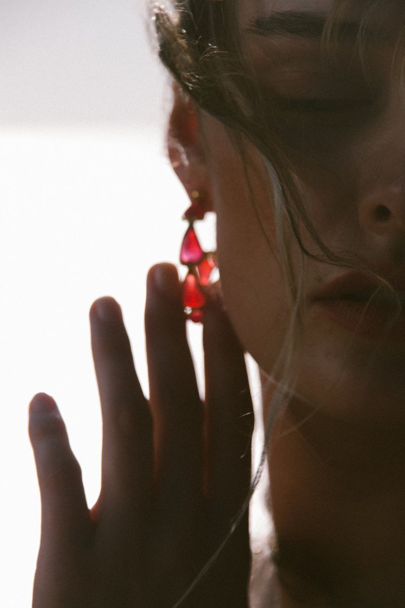 Close-up studio shot of a woman's face with bright, red teardrop earrings. Her fingers are softly touching her face.