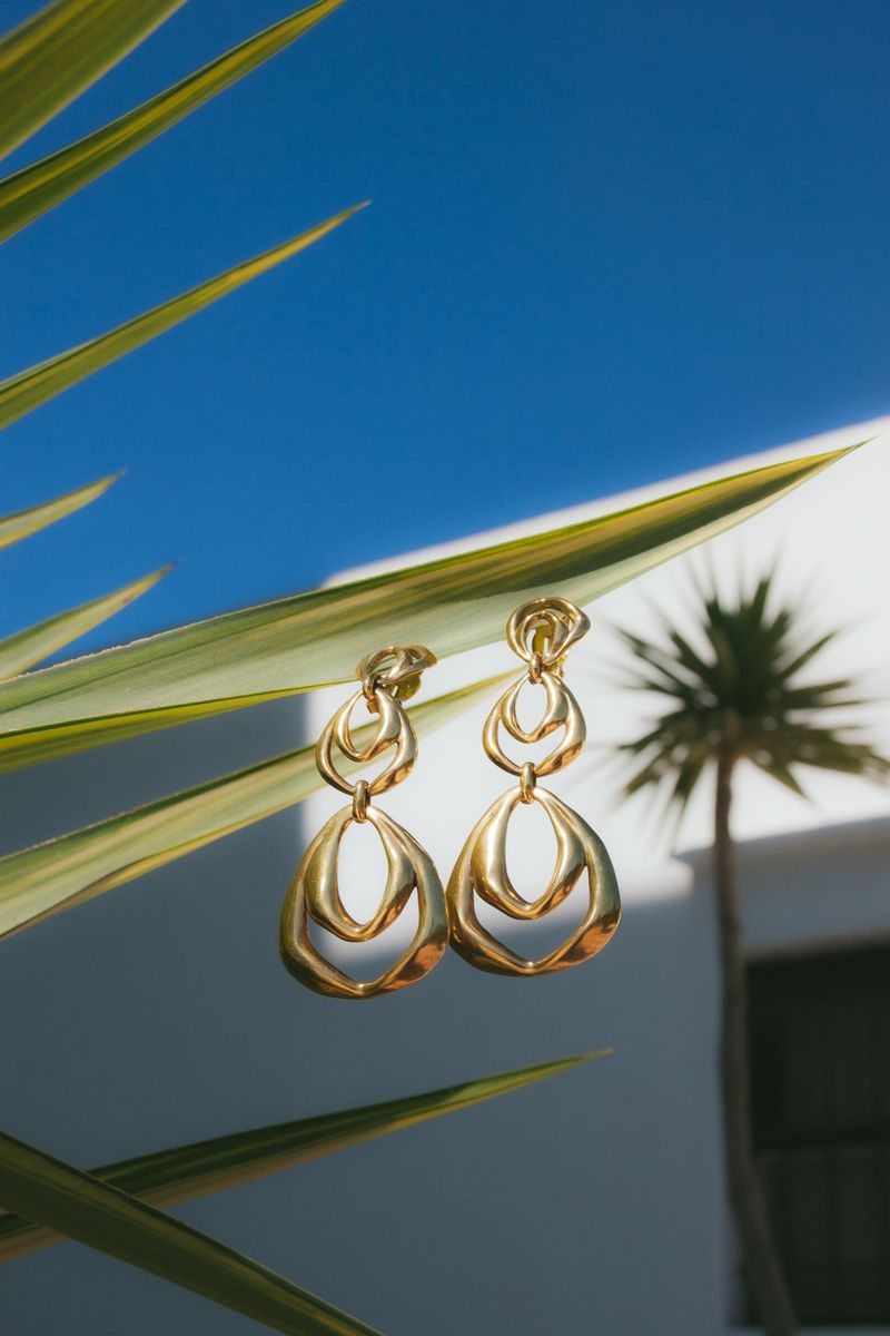 Hanging gold earrings with tropical leaf, blue sky, and building. Jewelry displayed against a bright outdoor backdrop.