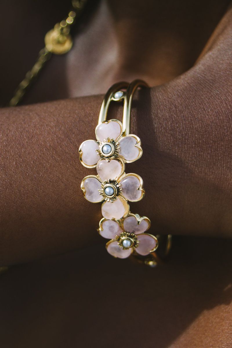 Close-up shot of a dark-skinned arm adorned with a gold cuff bracelet featuring light pink flower decorations with a pearl center.