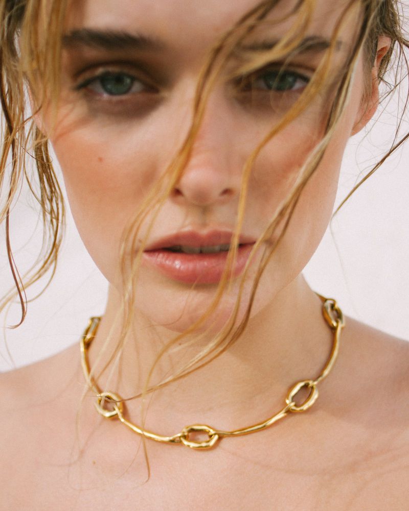 Close-up portrait of a woman with wispy strands of hair across her face wearing a textured gold link choker necklace.