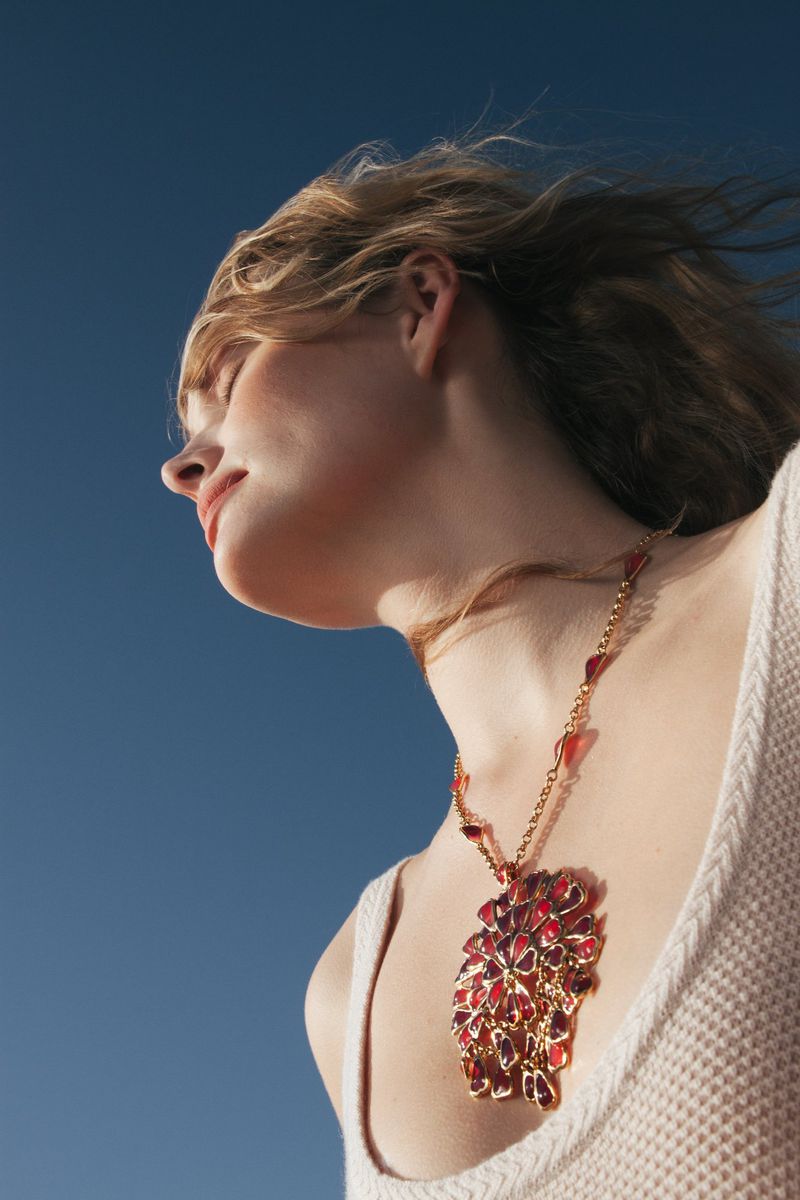A young woman with blonde hair poses in a low-angle shot, showcasing a large, elaborate red and gold necklace against a clear blue sky.