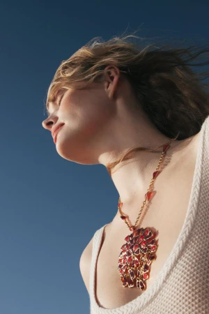 A young woman with blonde hair poses in a low-angle shot, showcasing a large, elaborate red and gold necklace against a clear blue sky.