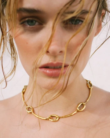 Close-up portrait of a woman with wispy strands of hair across her face wearing a textured gold link choker necklace.