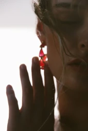 Close-up profile of a model wearing red statement drop earrings with a hand raised near her face against a bright background.