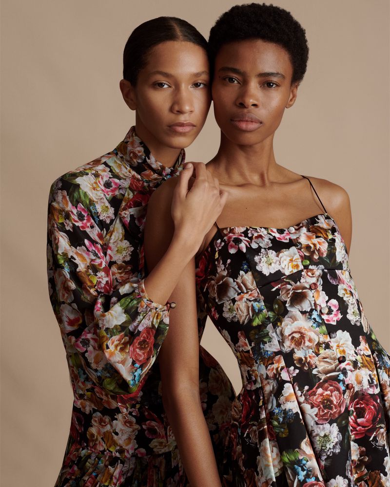 Two adult women pose in floral dresses against a neutral backdrop, showcasing fashion and style in a studio environment.