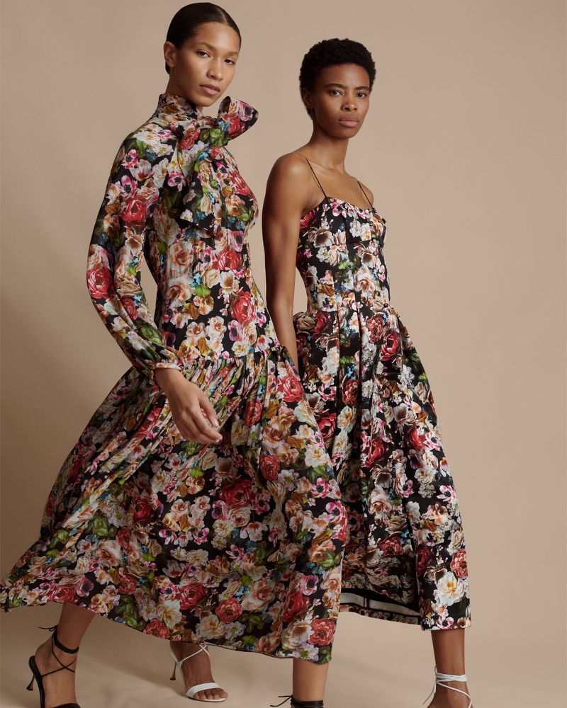 Two women wearing floral print dresses and heels pose against a neutral backdrop, showcasing fashion apparel.