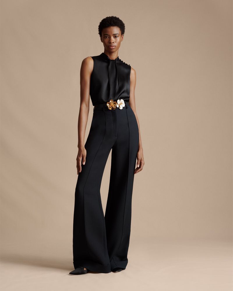 A woman stands in a studio setting wearing a black sleeveless blouse, wide-legged black pants, and a decorative gold and white belt.
