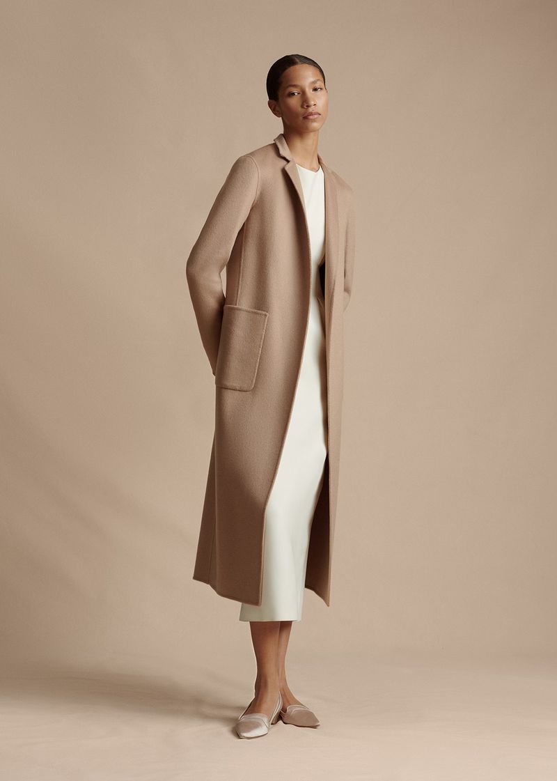 A woman models a long tan coat over a white dress with matching beige flats in a neutral, studio setting for a fashion shoot.