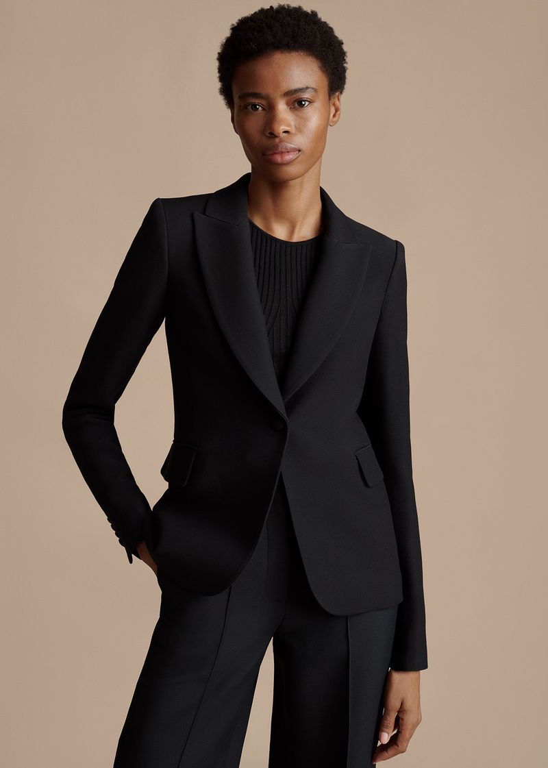 A woman with short hair models a black blazer and pants in a studio setting with a neutral backdrop, showcasing classic and professional style.