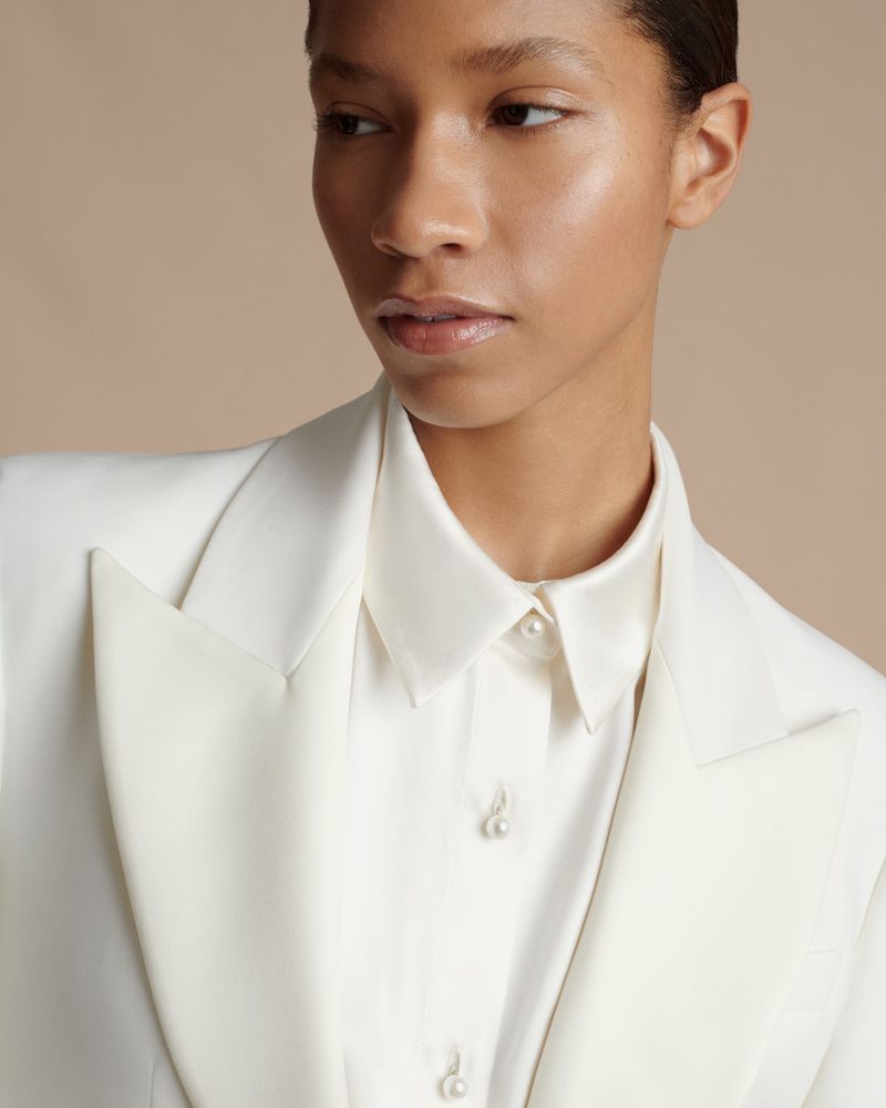 Close-up of an adult woman in a white blazer and pearl-buttoned shirt against a tan background in a studio-lit fashion portrait.