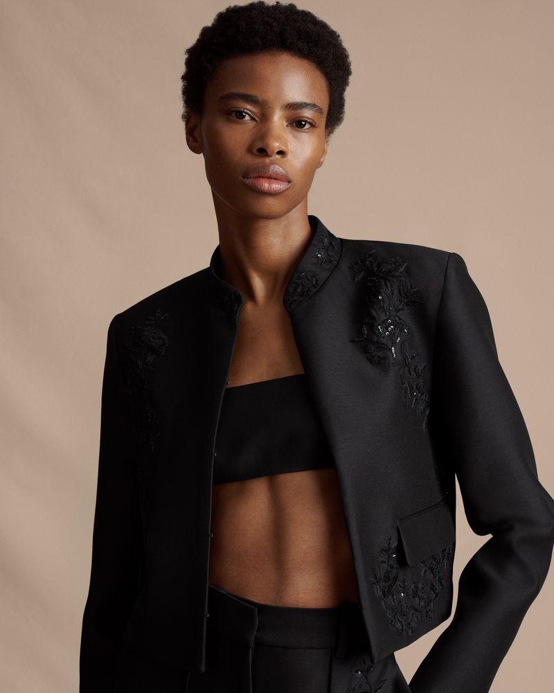 Elegant Black Woman in Embellished Blazer and Black Top, standing against a taupe backdrop. Studio lighting, portrait orientation.