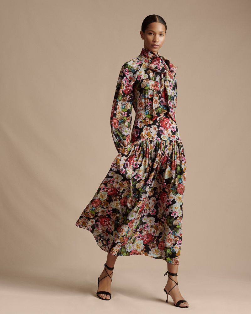 Elegant adult woman poses in a long-sleeve floral dress with strappy heels against a neutral background in a full-length fashion shot.