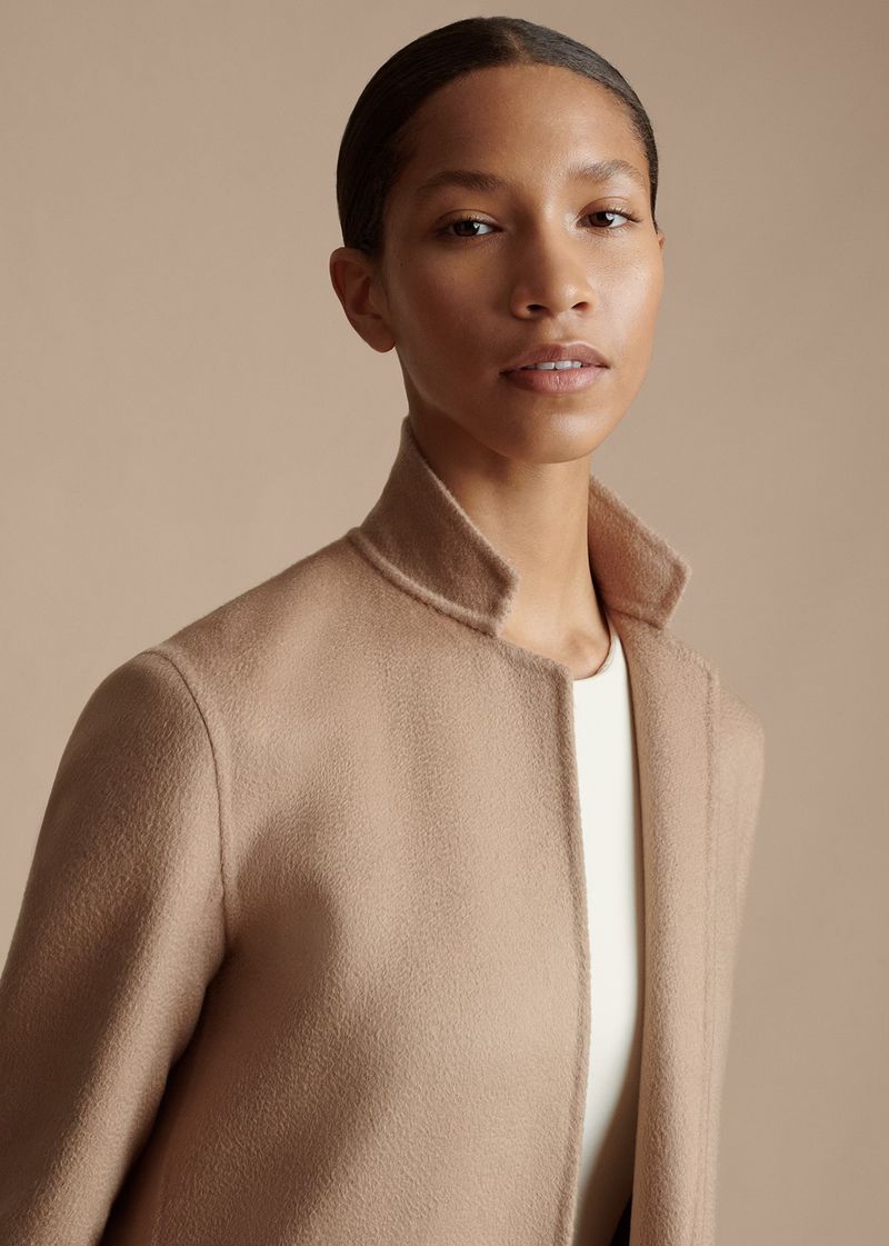 Close-up shot of a Black female adult posing indoors wearing a tan wool coat and white top against a neutral background in a portrait orientation.