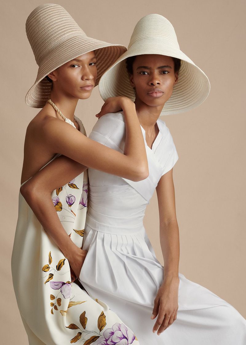 Two women pose in elegant, wide-brimmed hats and dresses with a neutral backdrop, fashion photography with soft lighting and a focus on style.