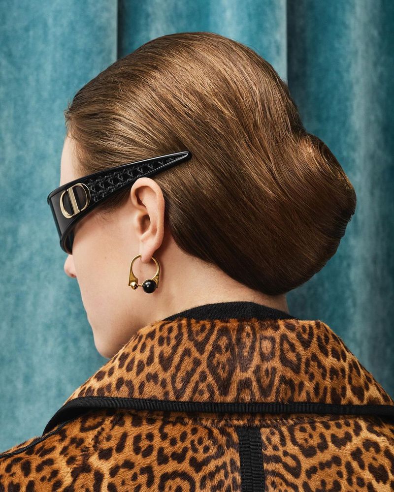 Elegant woman with brunette updo and statement earrings, wearing a leopard print coat and angular sunglasses with logo detail.