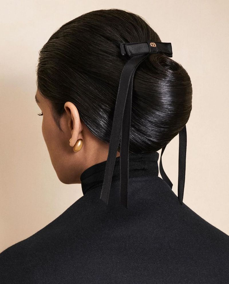 Woman's sleek updo with black ribbon bow, gold earrings, and a black turtleneck sweater against a neutral background, beauty shot.