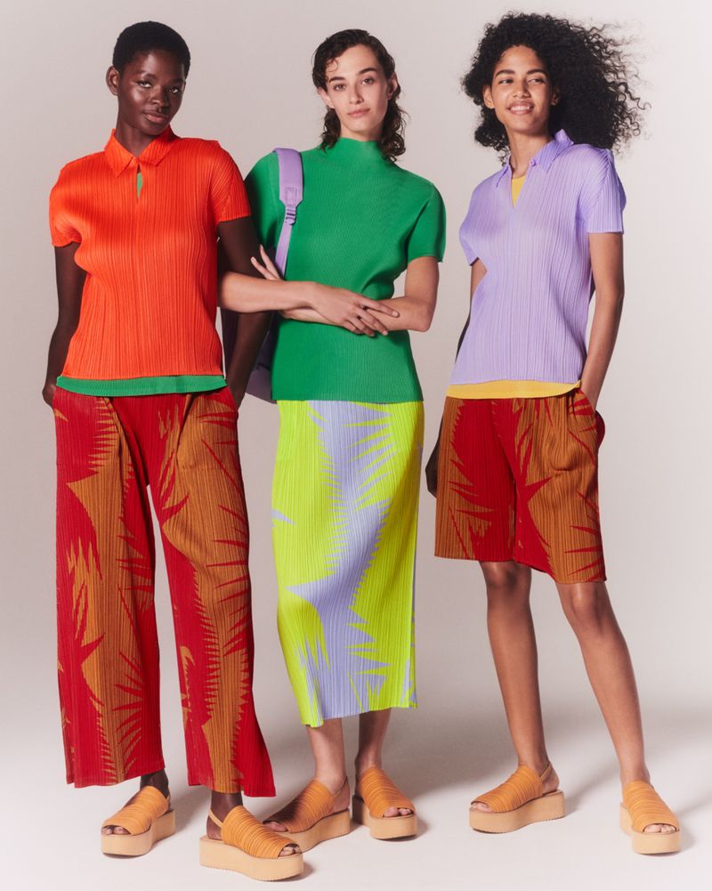 Three women are standing in a studio wearing orange, green, and lavender shirts with patterned skirts and pants, and matching platform sandals.