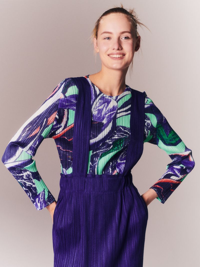 A fair-skinned woman smiles wearing a colorful patterned top and a purple overall dress, posed against a neutral studio backdrop.