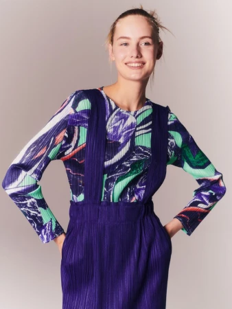A fair-skinned woman smiles wearing a colorful patterned top and a purple overall dress, posed against a neutral studio backdrop.