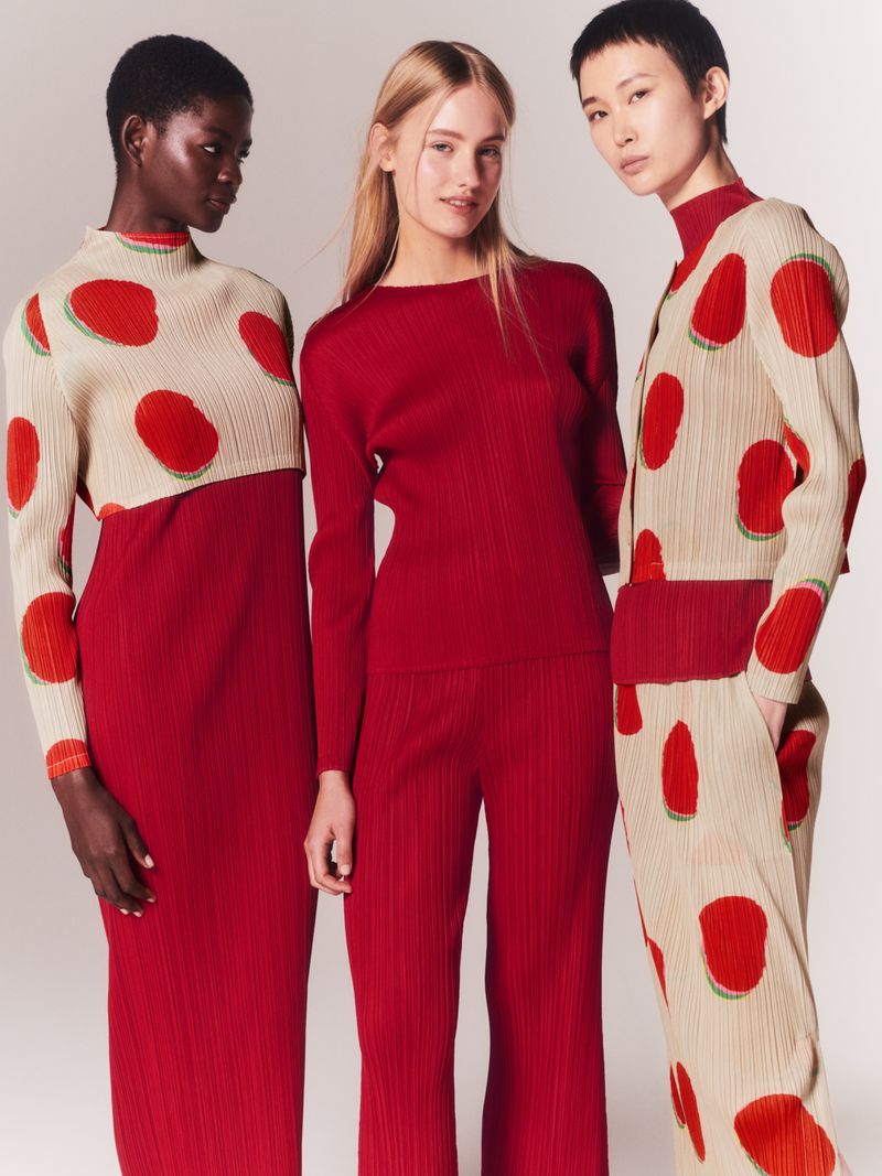 Three women in a studio wear matching pleated outfits in red, beige, and polka-dot patterned fabrics, posing against a grey backdrop.