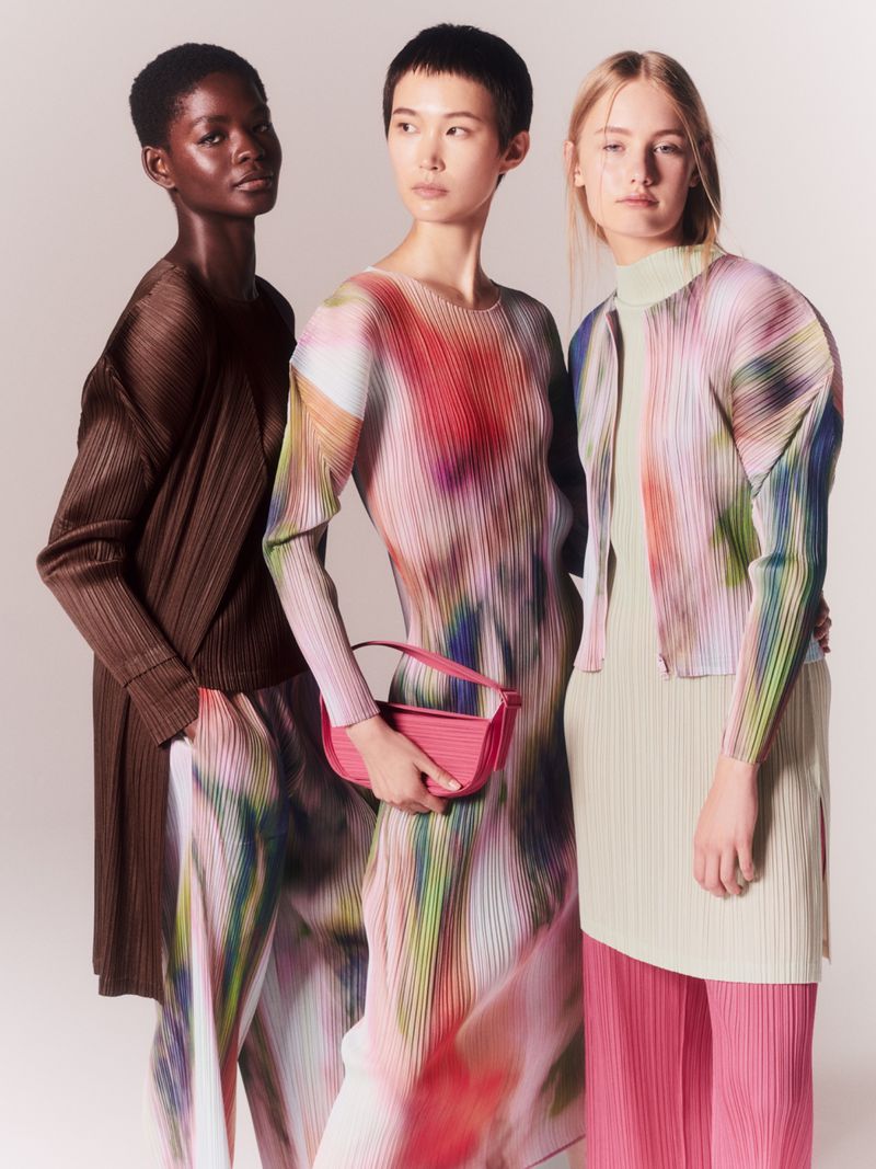 Three fashion models pose in plissé clothing with a muted, soft focus. A woman carries a small handbag with a short strap.