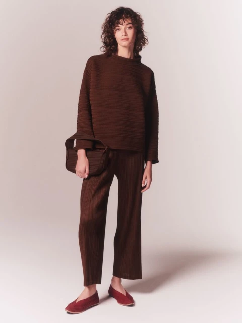 Full-length shot of a woman in a studio wearing a brown pleated outfit including a top, pants, a handbag, and flats.