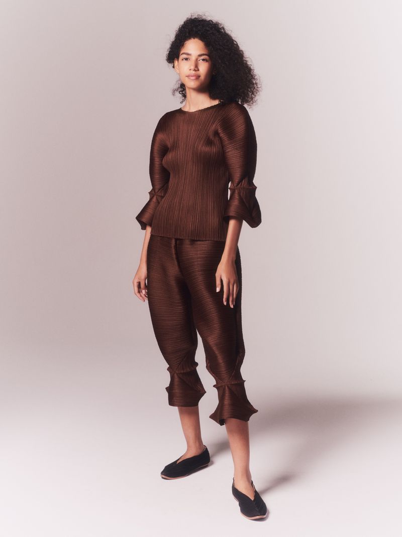 Fashion shot of a woman with curly hair wearing a brown pleated top and cropped pants with matching flats against a studio backdrop.