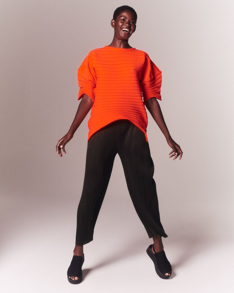 Smiling African-American woman poses in orange top and black pants in a studio setting, showcasing fashion and style with simple elegance.
