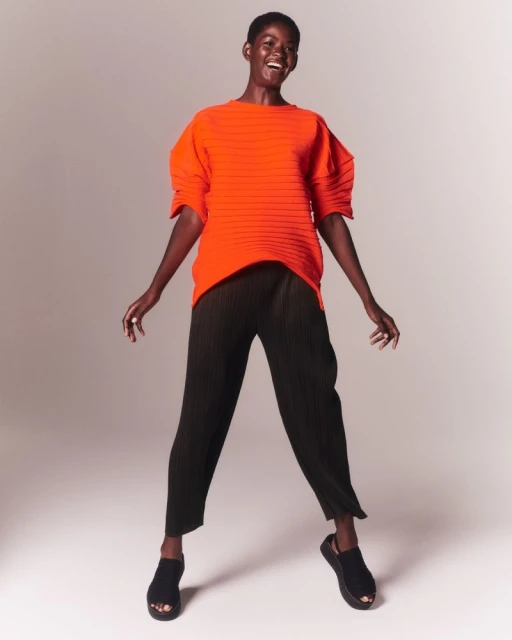Smiling African-American woman poses in orange top and black pants in a studio setting, showcasing fashion and style with simple elegance.