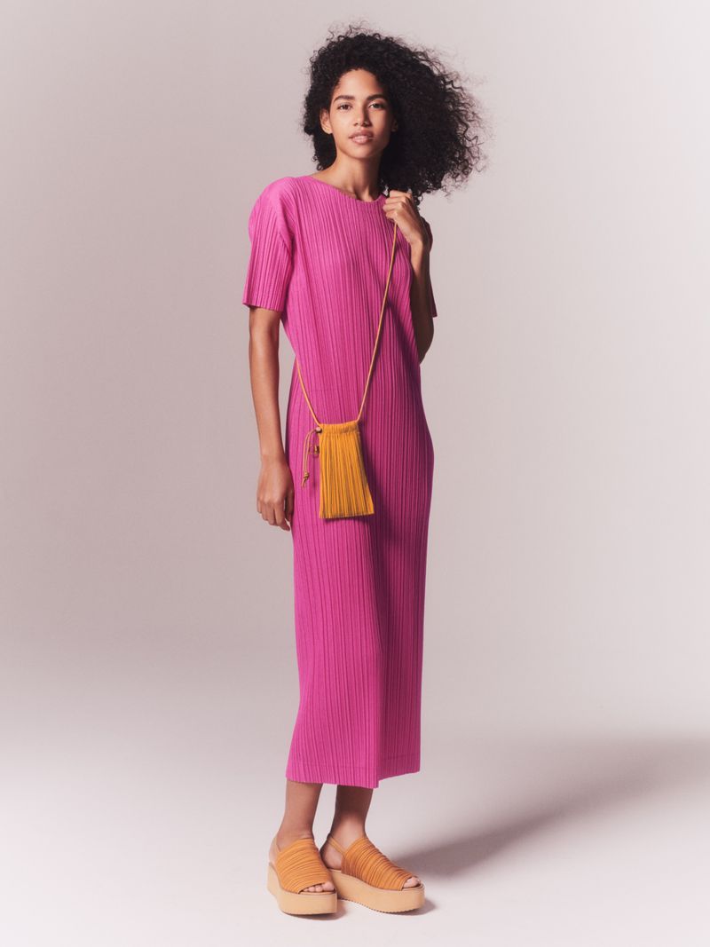 Full-length studio shot of a Black female model in a fuchsia dress, standing against a neutral background with a yellow purse.