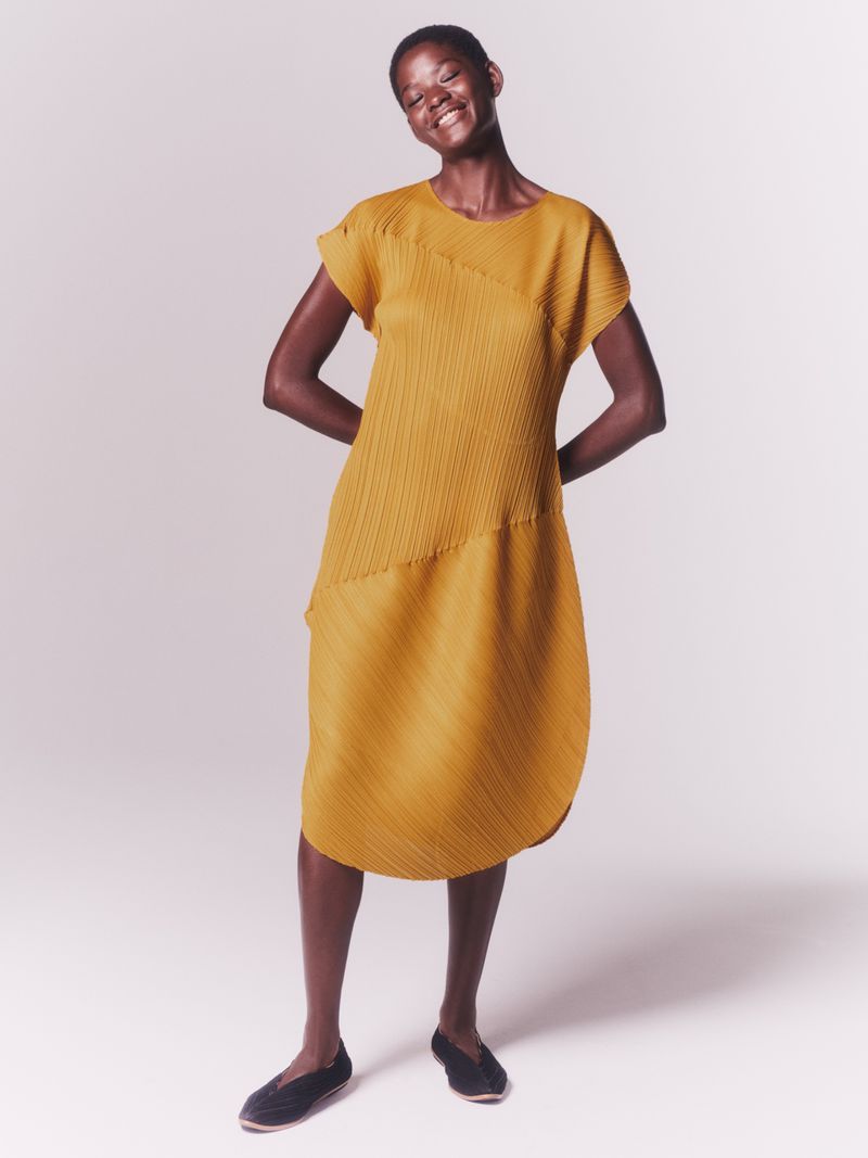 Dark-skinned woman in a honey-colored, pleated dress smiles in a studio shot, hands behind her back. Simple black flats.