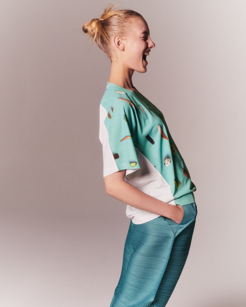 Side view of a smiling, fair-skinned young adult woman with blonde hair in a bun, wearing a light blue graphic tee and pleated pants.