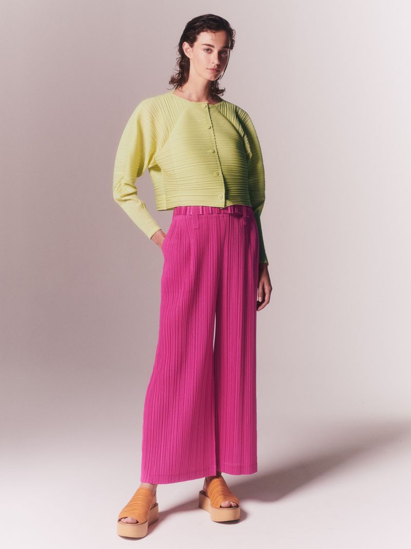 Fashion model poses in studio wearing a lime green ribbed jacket and magenta pleated pants, paired with tan platform sandals.