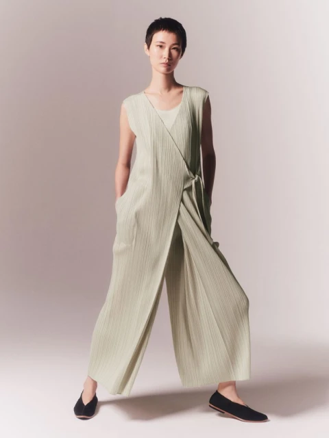 An adult woman poses in a flowing light green jumpsuit in a studio with gradient lighting, showcasing the garment's draped design.
