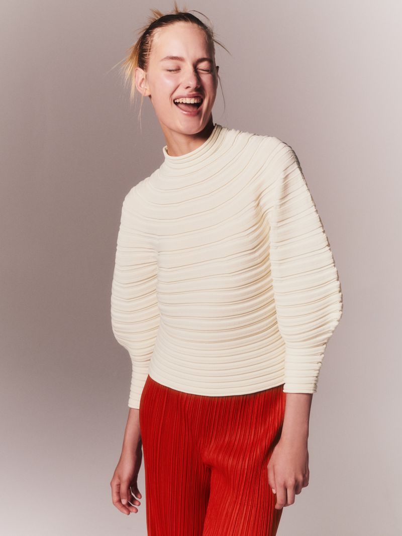 A fair-skinned woman smiles, wearing a cream-colored ribbed top and red pleated pants, against a light grey studio backdrop in a medium shot.