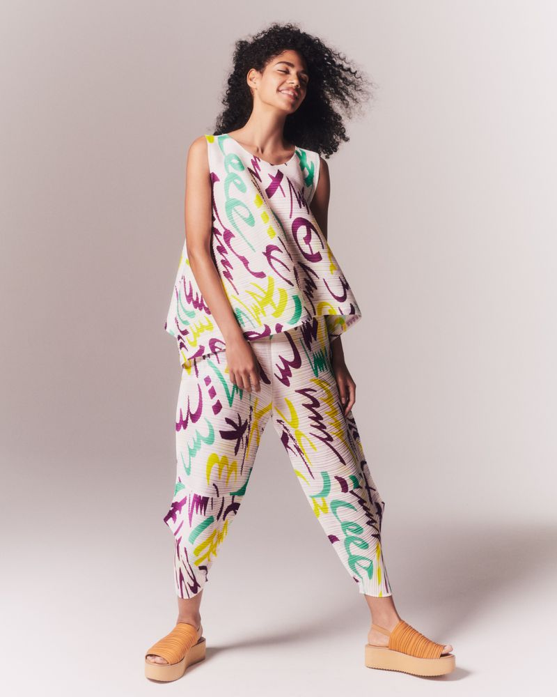 An adult woman in a white sleeveless top and pants with colorful abstract designs, standing in a bright photo studio, smiling slightly.