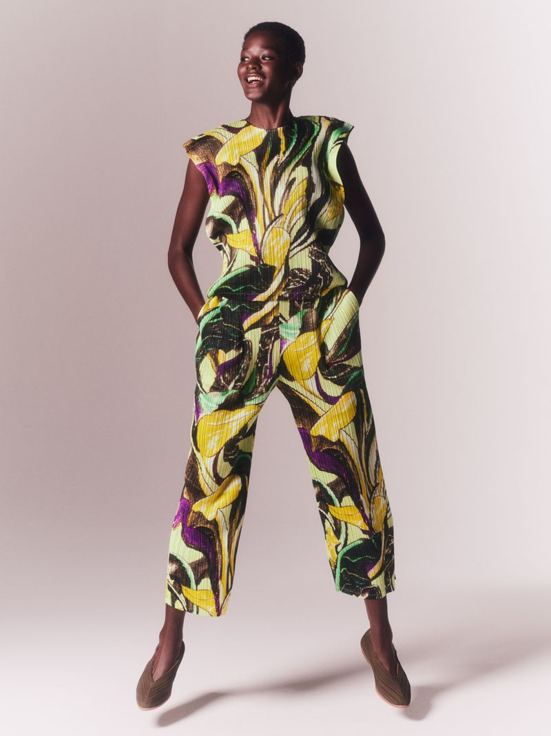 Smiling Black female fashion model in a bright floral top and culottes, mid-air in a photo studio, hands in pockets, looking off-camera.