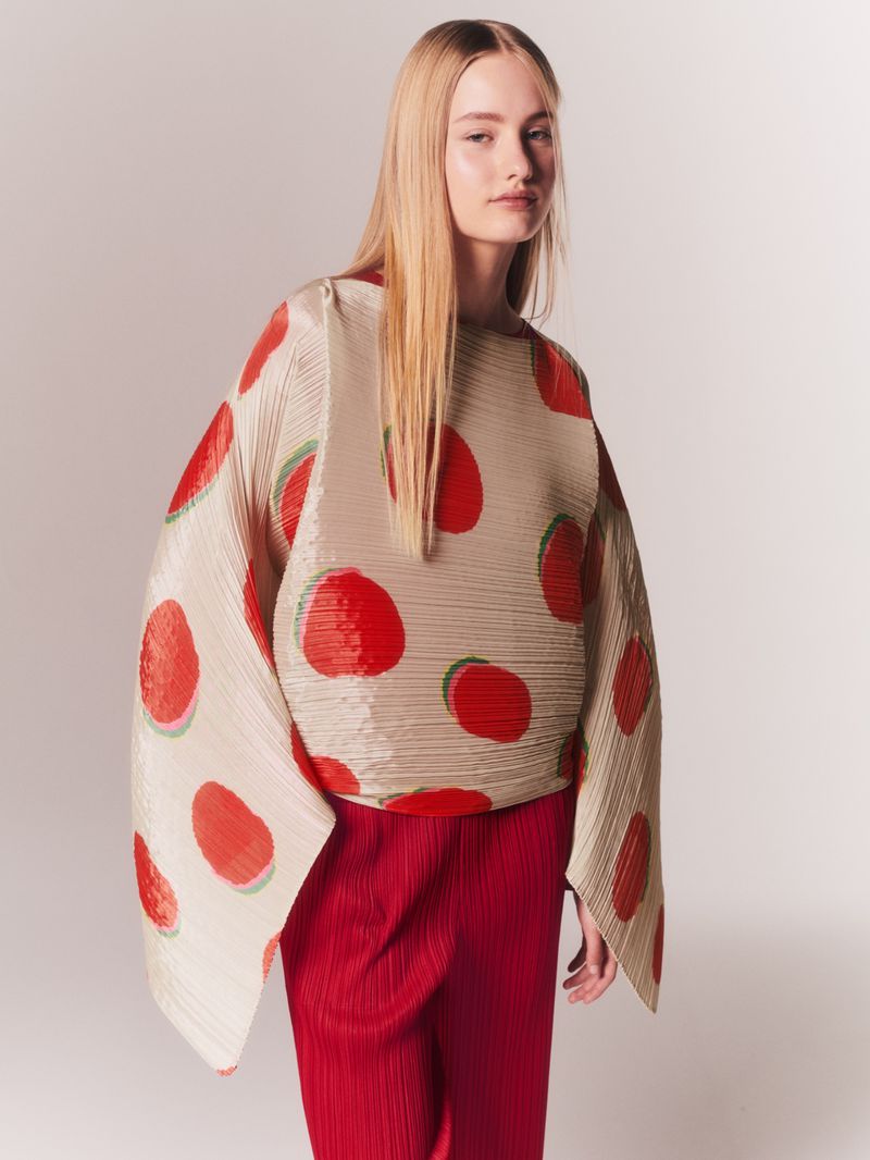 Fashion shot of a fair-skinned woman with long blonde hair wearing a beige and red polka dot blouse and red pants against a white background.