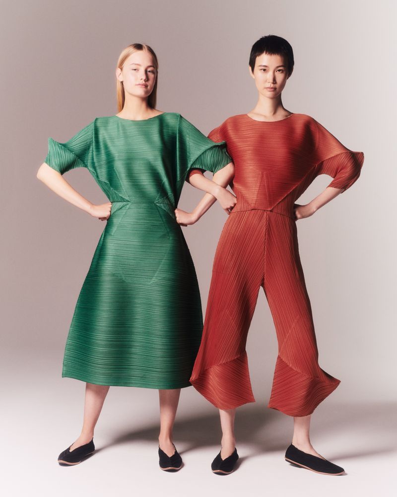 Two women pose indoors wearing pleated dresses in green and reddish-brown, with short hair and black shoes, against a neutral background.
