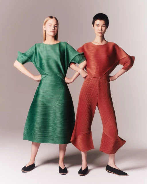 Two women pose indoors wearing pleated dresses in green and reddish-brown, with short hair and black shoes, against a neutral background.