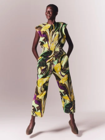 Model wearing a yellow and green pleated patterned sleeveless jumpsuit and brown flat shoes against a white studio background.