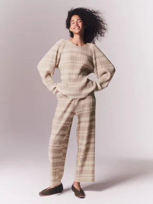 Model with dark curly hair wearing a cream pleated top and trousers against a neutral background for Issey Miyake Pleats Please S/S 24.