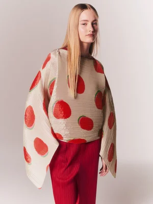 Model wearing an oversized pleated cream top with red circular patterns and matching red pleated trousers against a pale studio backdrop.
