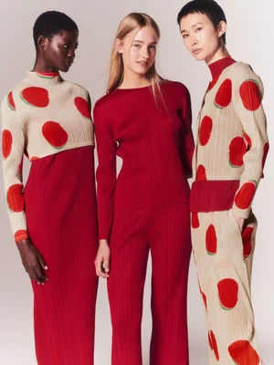 Three female models standing closely in red and beige patterned pleated garments against a neutral studio background.