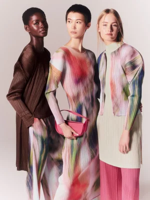 Three models in colorful textured pleated outfits and a pink handbag standing together against a studio background.