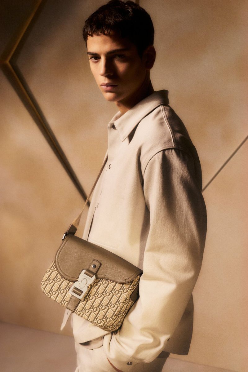 A young man in a light-beige jacket poses indoors with a designer beige saddle bag, showcasing a fashion-forward, monochromatic look.