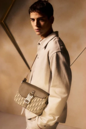 A young man in a light-beige jacket poses indoors with a designer beige saddle bag, showcasing a fashion-forward, monochromatic look.