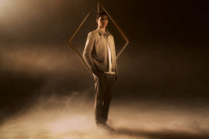Model Tobias Baez wearing a beige tailored jacket and trousers standing before a suspended gold diamond frame amidst smoke.