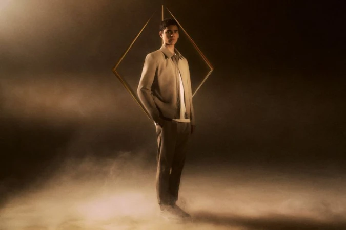 Model Tobias Baez wearing a beige tailored jacket and trousers standing before a suspended gold diamond frame amidst smoke.
