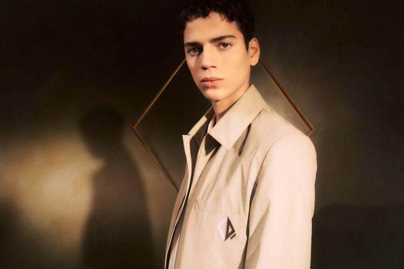 Portrait of a young adult male in a beige jacket, posing in front of a framed diamond shape, with a dark greenish-brown background.